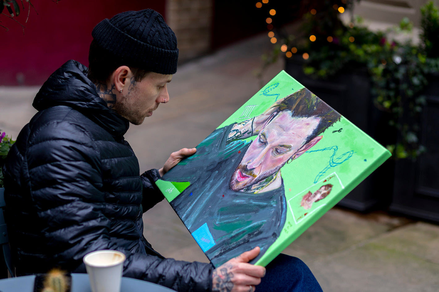  Ben from The New Normal Charity sitting on a chair next to a table with a cup of coffee, looking at his commissioned portrait with a focused expression.